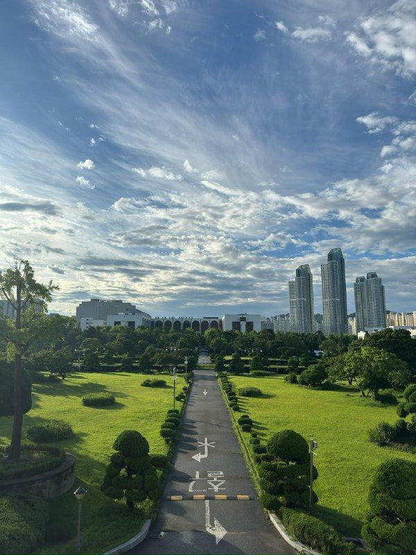 A park path leading towards city towers under a wide sky of clouds