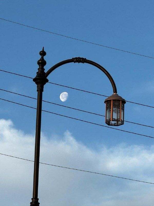 An ornate street lamp and power lines against blue sky with a daytime moon