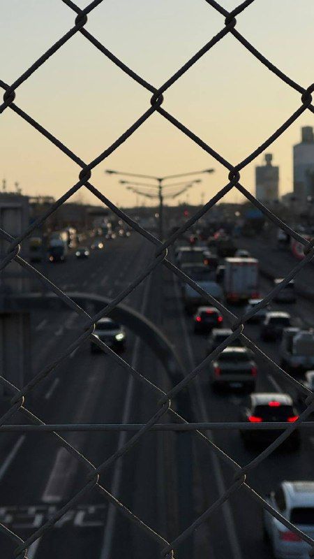 Highway traffic at dusk seen through a chain-link fence, city towers on the horizon