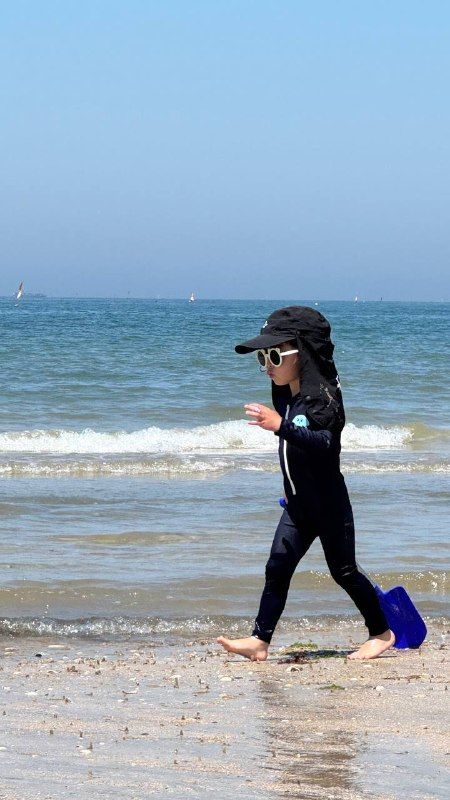 A child in a cap and wetsuit walking along the shoreline with a spade