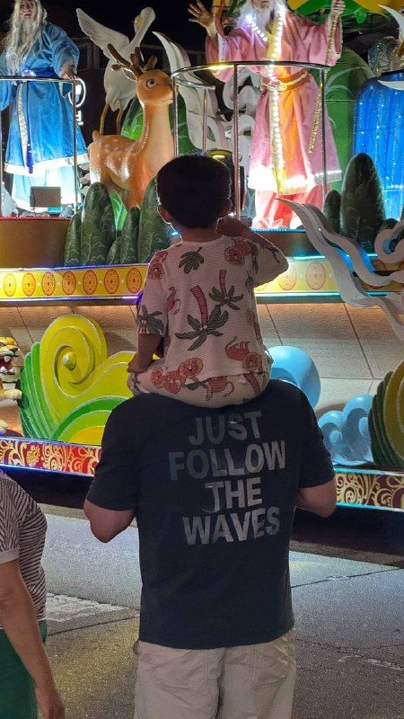 A boy in a "just follow the waves" shirt on his father’s shoulders before a lantern float