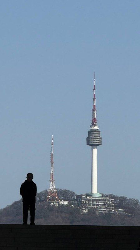 A lone figure silhouetted beside N Seoul Tower in flat blue haze