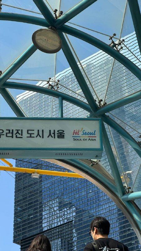 A glass-roofed bus shelter with a "Hi Seoul" sign beside office towers