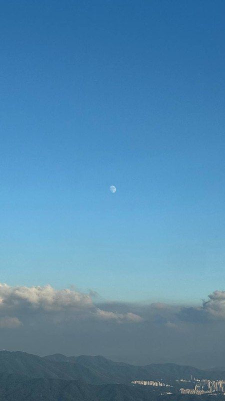 A daytime moon above a band of cloud over blue hills
