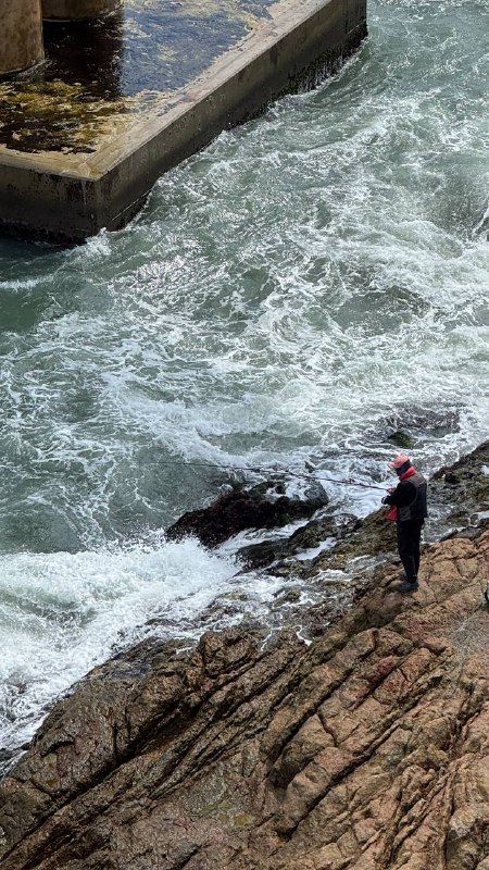 A lone figure on dark rocks as waves crash white below
